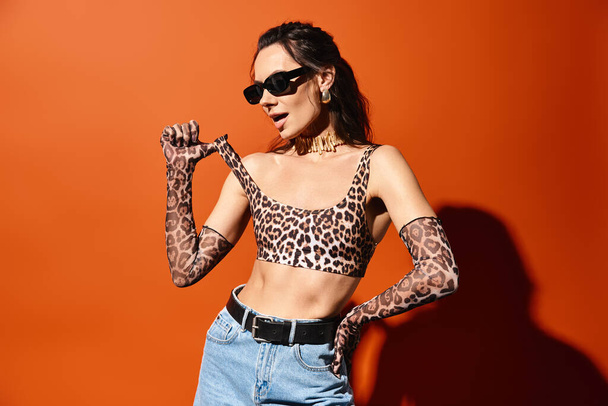Une femme à la mode porte en toute confiance un haut imprimé léopard et un jean, accessoirisé avec des lunettes de soleil, sur fond de studio orange. - Photo, image