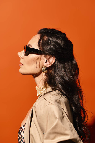 Μια κομψή γυναίκα με γυαλιά ηλίου θέτει με σιγουριά σε ένα μοντέρνο σακάκι σε ένα πορτοκαλί φόντο. - Φωτογραφία, εικόνα