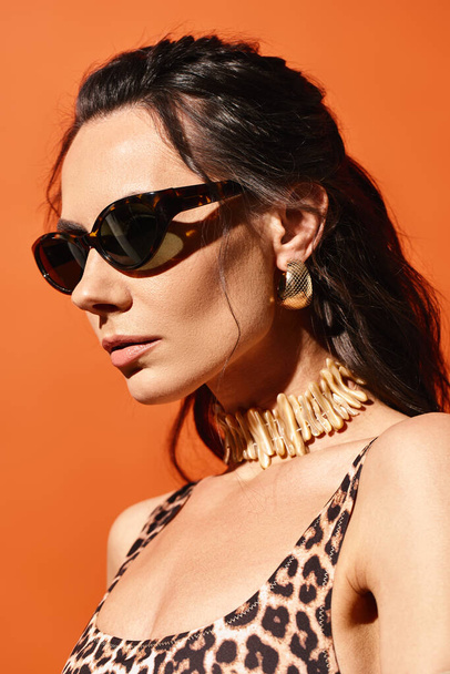 Una mujer elegante con gafas de sol posa en una parte superior de impresión de leopardo sobre un fondo de estudio naranja, exudando vibraciones de moda de verano. - Foto, imagen