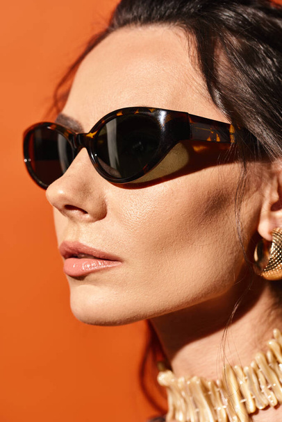 A fashionable woman exudes summer vibes with sunglasses and a gold necklace against an orange backdrop. - Photo, Image