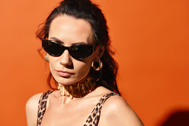 Une femme à la mode portant des lunettes de soleil et un haut imprimé léopard pose en toute confiance dans un studio sur un fond orange. - Photo, image