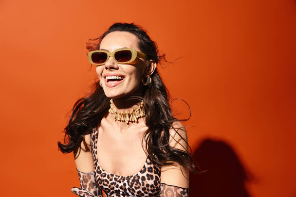 Μια μοντέρνα γυναίκα αποπνέει εμπιστοσύνη σε ένα λεοπάρ φόρεμα εκτύπωσης και κομψά γυαλιά ηλίου σε ένα πορτοκαλί φόντο στούντιο. - Φωτογραφία, εικόνα
