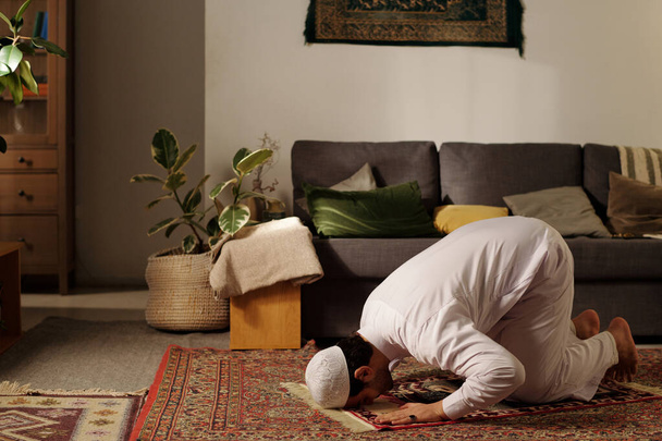 Młody człowiek z Bliskiego Wschodu ćwiczący namaz na dywanie modlitewnym sujud w salonie w domu, kopiowanie przestrzeni - Zdjęcie, obraz