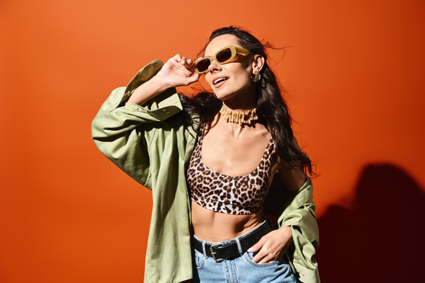 Uma mulher elegante vestindo um top de impressão de leopardo e jeans, exalando confiança e moda de verão em um fundo de estúdio laranja. - Foto, Imagem