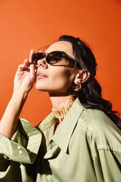 Une femme élégante avec des lunettes de soleil et une chemise verte pose sur un fond de studio orange, respirant les vibrations de la mode estivale. - Photo, image