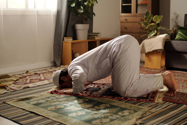 Młody muzułmanin w białych ubraniach robi sajdah ćwicząc namaz na dywanie modlitewnym w salonie w domu - Zdjęcie, obraz
