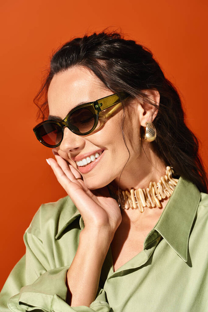 Une femme élégante avec des lunettes de soleil pose en toute confiance dans une chemise verte sur fond de studio orange. - Photo, image