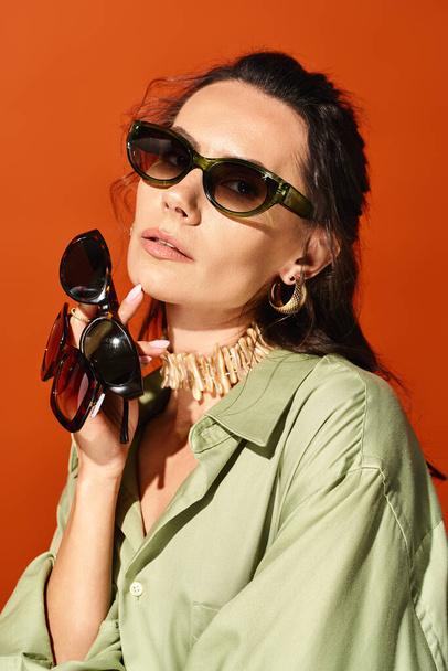 Yeşil gömlekli şık bir kadın, stüdyoda turuncu bir arka planda bir güneş gözlüğü takıyor ve yaz modası havası yayıyor.. - Fotoğraf, Görsel