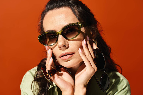 Стильная женщина в солнцезащитных очках и зеленой рубашке позирует в студии с ярким оранжевым фоном, источая летние модные флюиды. - Фото, изображение