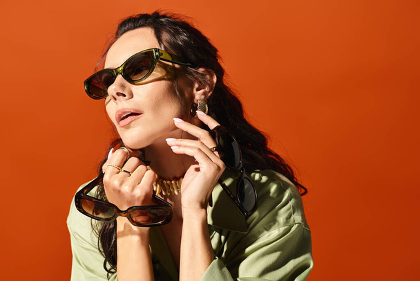 Modna kobieta pewnie pozuje w studio w okularach przeciwsłonecznych i zielonej koszuli, emanując latem wibracjami na pomarańczowym tle. - Zdjęcie, obraz