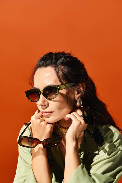 Стильна жінка в сонцезахисних окулярах вражає впевнену позу в студії на яскравому помаранчевому фоні, виключаючи літній час моди. - Фото, зображення