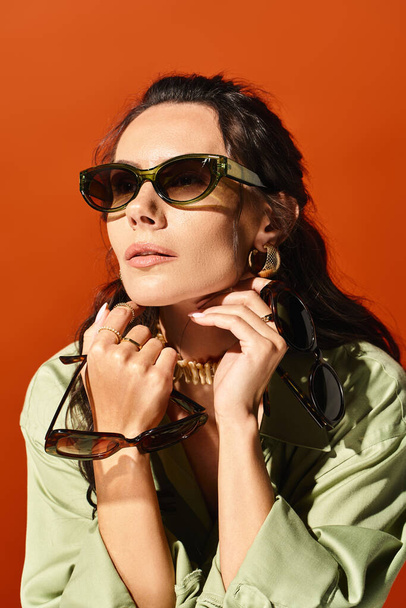 Una mujer elegante con gafas de sol posa en una camisa verde en un estudio sobre un fondo naranja, exudando vibraciones de moda de verano. - Foto, Imagen