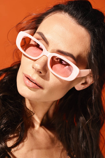 Модная женщина с длинными волосами в розовых солнцезащитных очках, излучающих летние вибрации в студии на оранжевом фоне. - Фото, изображение