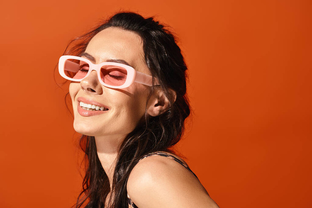 Стильная женщина в розовых солнцезащитных очках улыбается в студии на оранжевом фоне, излучая летние модные флюиды. - Фото, изображение