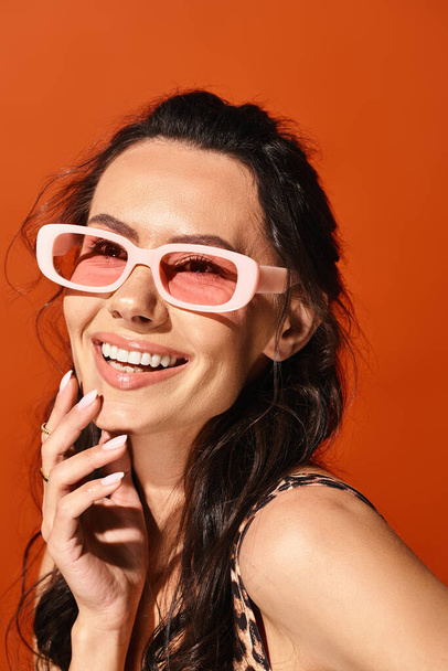 A stylish woman is striking a pose while wearing pink sunglasses against an orange studio backdrop. - Foto, afbeelding