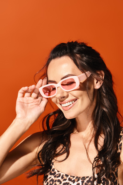 Uma mulher elegante exala confiança em óculos de sol rosa e um top de impressão de leopardo contra um fundo laranja. - Foto, Imagem