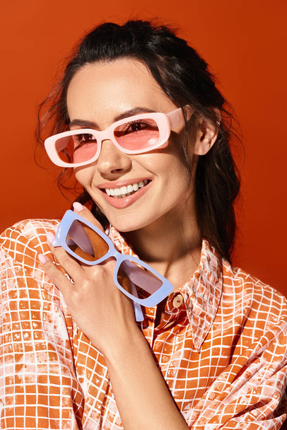 Una mujer elegante con gafas de sol de color rosa y una camisa a cuadros sobre un fondo naranja. - Foto, imagen