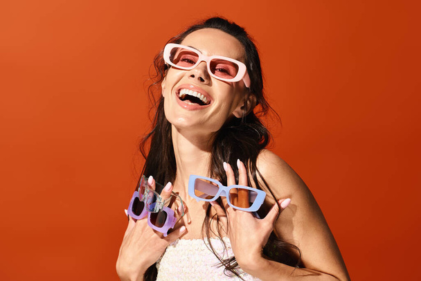 Una mujer impresionante exuda felicidad mientras sonríe hacia la cámara con elegantes gafas de sol, contra un vibrante fondo de estudio naranja. - Foto, imagen