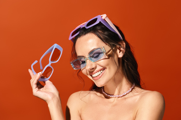 Una mujer con estilo en gafas de sol sosteniendo un par de gafas en un estudio sobre un fondo naranja, mostrando la moda de verano. - Foto, imagen