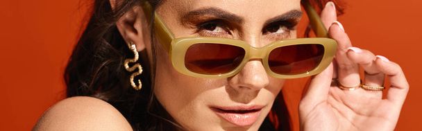 Modaya uygun bir kadın, turuncu bir arka planda parlak sarı güneş gözlüklerini sallayarak, yaz mevsiminde stüdyoda titreşim yayar.. - Fotoğraf, Görsel