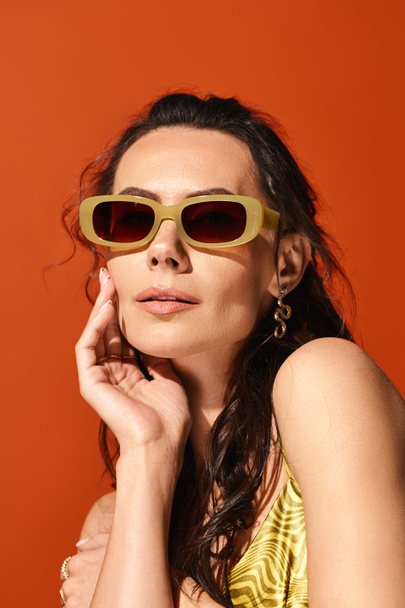 Una mujer con estilo en un vestido amarillo vibrante y gafas de sol elegantes posa elegantemente sobre un telón de fondo naranja, exudando vibraciones de verano. - Foto, imagen