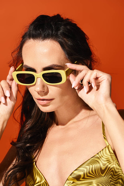 Una mujer elegante con una sonrisa radiante lleva un top amarillo vibrante y gafas de sol de moda contra un fondo naranja audaz. - Foto, Imagen