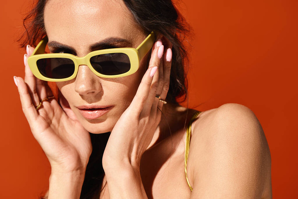 Una mujer de moda con gafas de sol amarillas, las manos en la cara, en el estudio sobre fondo naranja. - Foto, imagen
