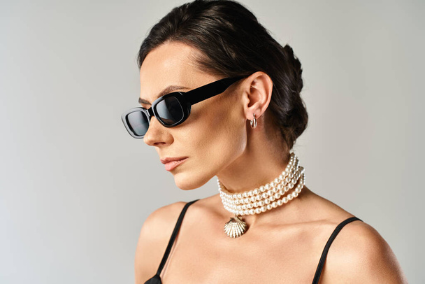 Modna kobieta w stylowych okularach przeciwsłonecznych i eleganckim naszyjniku pozuje w studio na szarym tle. - Zdjęcie, obraz