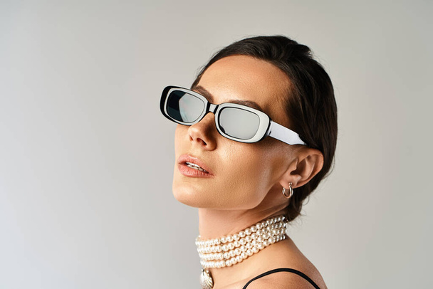 Una mujer elegante con gafas de sol y un collar de perlas emana confianza en un estudio sobre un fondo gris. - Foto, imagen