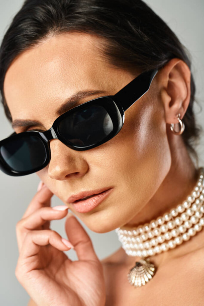 A fashionable woman exudes sophistication in black sunglasses and a pearl necklace against a neutral backdrop. - Foto, imagen