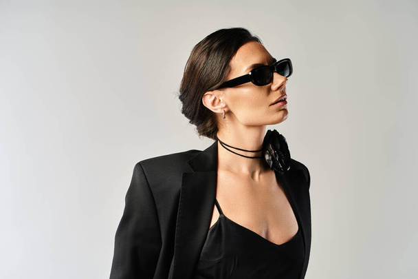 Eine stilvolle und geheimnisvolle Frau trägt in einem Studio vor neutralem grauen Hintergrund einen eleganten schwarzen Anzug und eine trendige Sonnenbrille. - Foto, Bild