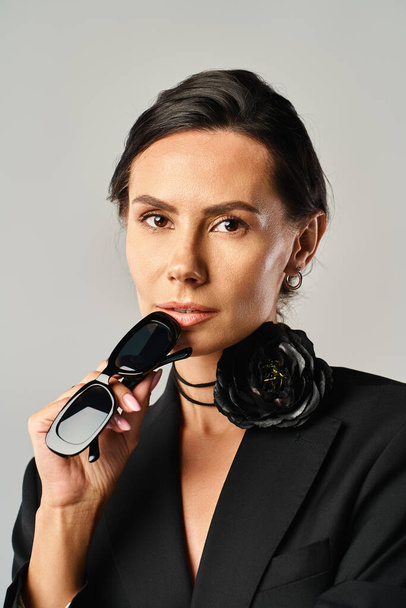 Eine stilvolle Frau im schwarzen Anzug hält eine Sonnenbrille in der Hand und präsentiert Eleganz und Raffinesse in einem Studio vor grauem Hintergrund. - Foto, Bild