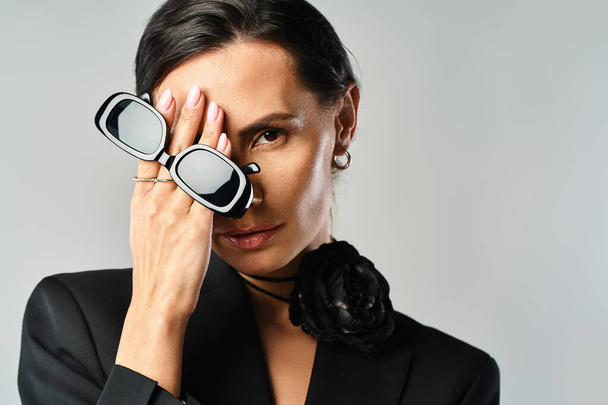 Eine stilvolle Frau im schwarzen Anzug hält selbstbewusst eine Brille in einem Studio-Setting mit grauem Hintergrund. - Foto, Bild