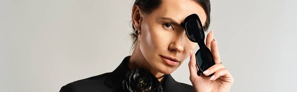 Eine stilvolle Frau im Anzug hält selbstbewusst eine schwarze Sonnenbrille in einem Studio-Setting auf grau - Foto, Bild