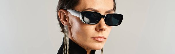 Una mujer de moda que luce gafas de sol y una camisa negra posa con confianza en un estudio sobre un fondo gris. - Foto, imagen