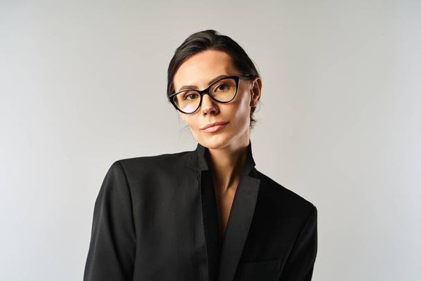 Uma mulher elegante usando óculos e uma jaqueta preta posa em um estúdio contra um fundo cinza. - Foto, Imagem