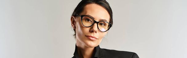Una mujer de moda exuda sofisticación en una camisa negra, complementada con gafas de sol elegantes, contra un fondo de estudio neutral. - Foto, imagen