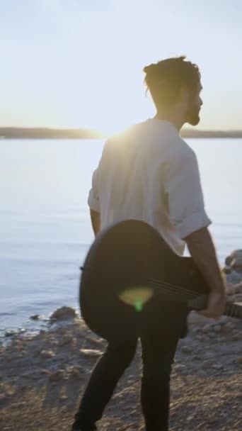 Hipster hombre con la guitarra disfrutar de la puesta de sol en la costa del lago caminando en la playa de piedra contra el sol - cámara lenta vista trasera - Vertical FullHD video - Imágenes, Vídeo