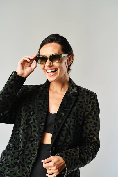 A fashionable woman in a black jacket and sunglasses poses in a studio setting against a grey background. - Photo, Image