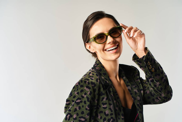 Egy elegáns nő pózol leopárdmintás dzsekiben és divatos napszemüvegben, szürke stúdió háttérrel.. - Fotó, kép