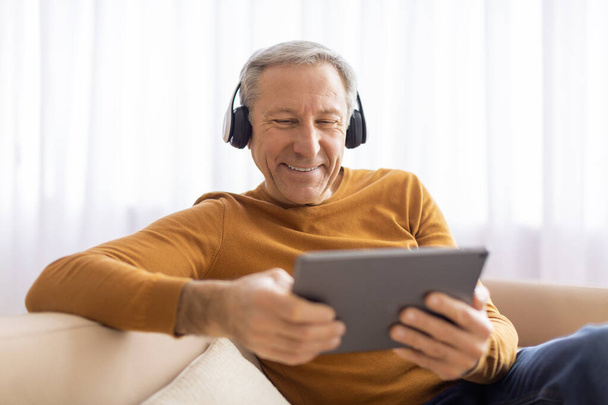 Starszy mężczyzna wygląda na rozbawionego, gdy używa tabletu ze słuchawkami, siedząc wygodnie w ciepłym, przytulnym pokoju. - Zdjęcie, obraz