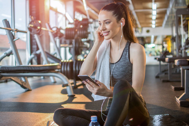 Young fit woman with earphones listening to music on her smart phone and taking a rest on fitness mat after gym workout - Photo, Image