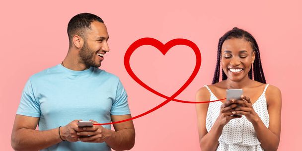 An engaging man in blue t-shirt looks at a joyful woman in a white polka dot dress, both using smartphones with a red heart graphic implying a connection between them, set against a pink background - Photo, Image