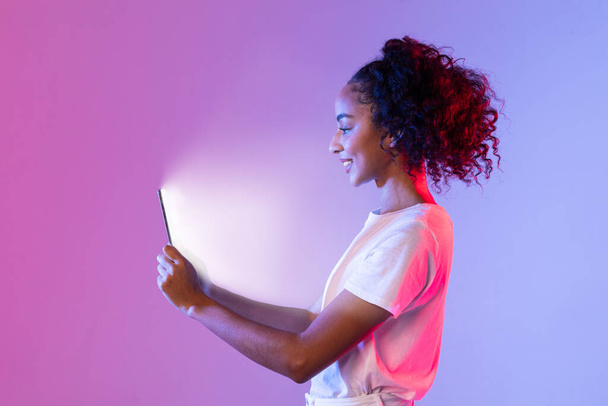Boční profil šťastné černošky s kudrnatými vlasy, dívající se na tablet, jehož obrazovka osvětluje její tvář, proti fialové a růžové gradient neonové pozadí - Fotografie, Obrázek