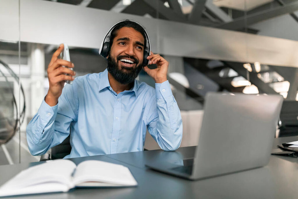 Modern ofis ortamında kulaklık takan ve akıllı telefon tutan neşeli bir profesyonel uzaktan iletişimi gösteriyor. - Fotoğraf, Görsel