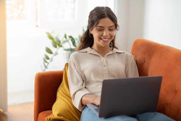 Comunicação online. Sorrindo jovem fêmea usando laptop enquanto sentado no sofá em casa, feliz mulher milenar digitando no computador enquanto relaxa na acolhedora sala de estar interior, espaço de cópia - Foto, Imagem