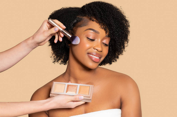 Sorrindo jovem mulher negra gosta de ter iluminador escovado em suas maçãs do rosto, artista segurando paleta, incorporando o prazer de aplicação de maquiagem - Foto, Imagem