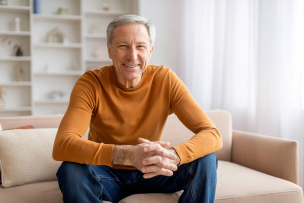 Pozitív jóképű, ősz hajú, idősebb férfi otthon ül a kanapén, és mosolyog a kamerába. Boldog nagyapa élvezi a hétvégét, hangulatos nappali belső, másolás tér - Fotó, kép