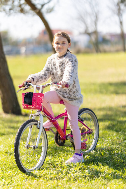 Ένα νεαρό κορίτσι κάθεται σε ένα ροζ ποδήλατο σε ένα πάρκο. Φοράει ροζ πουλόβερ και ροζ παντελόνι. - Φωτογραφία, εικόνα