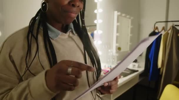 Közepes felvétel fiatal fonott fekete nő olvasás és felülvizsgálata anyagot írt papírlapokra, miközben készül stand up koncert a színfalak mögött szobában - Felvétel, videó